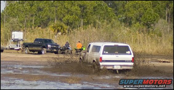 White Ford Bronco driving through mud, rear view. "FORD" visible on tailgate
