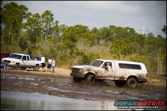 White Ford Bronco with camper shell driving through mud, 3q-front view