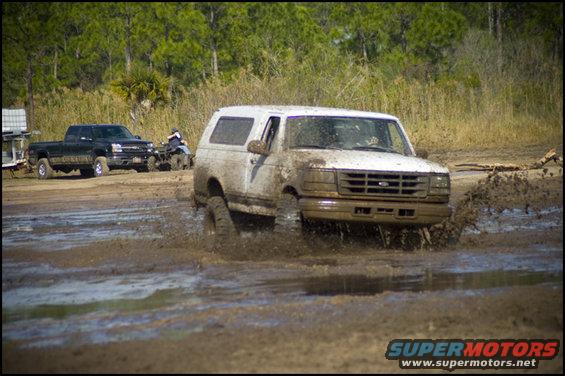 White Ford Bronco driving through mud, black pickup and ATV in background