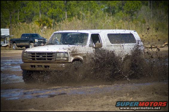 White SUV splashing mud, front view. Another black pickup truck is visible in the background