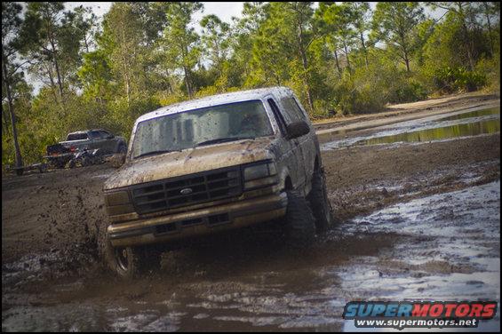 Muddy Ford Bronco driving through a muddy track with another truck in the background