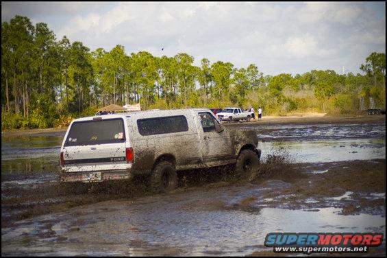 White Ford Bronco driving through mud, splashing water. Mud covers the vehicle