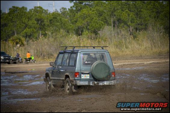 Gray Mitsubishi Montero driving through mud, rear view. License plate visible