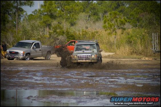 Two trucks and an SUV driving through mud, kicking up water and dirt