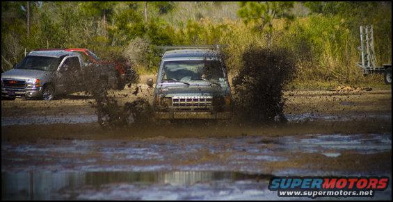 Two trucks driving through mud, one is kicking up a lot of spray