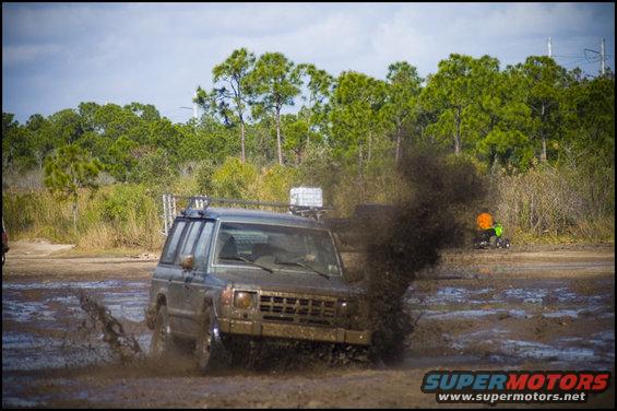 Mud-splattered SUV with roof rack driving through a muddy track, kicking up water