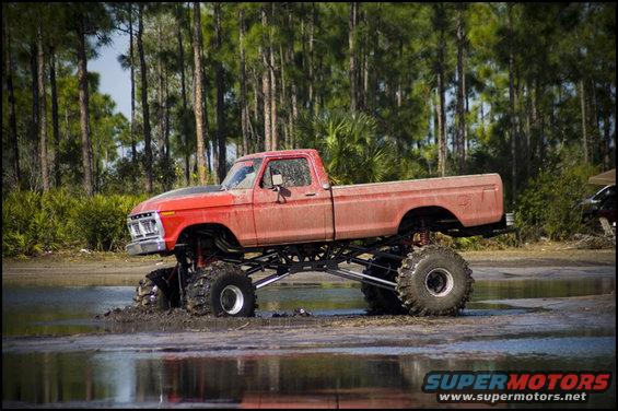 red lifted monster truck driving through mud and water, trees in background