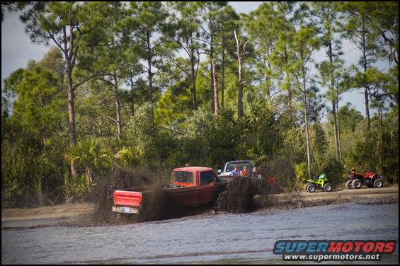 Red pickup truck and white SUV churning mud, two ATVs parked nearby. Trees and water in background