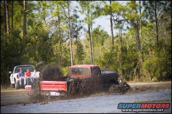 Red and black pickup truck kicking up mud, white pickup truck with people watching in background