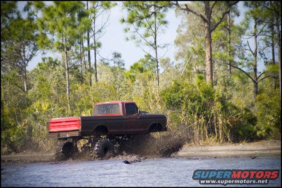 Red and brown Ford F-Series pickup truck with large tires driving through mud