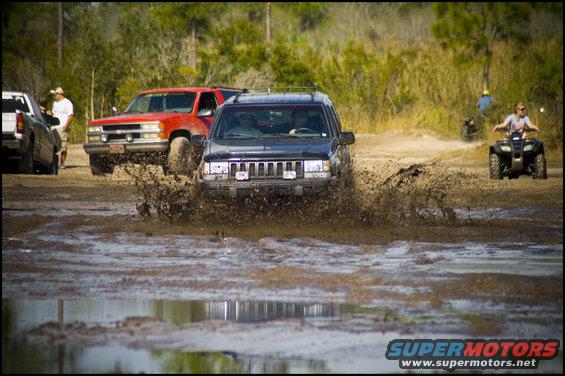 Jeep Grand Cherokee splashing through mud, red truck behind it, ATVs in background