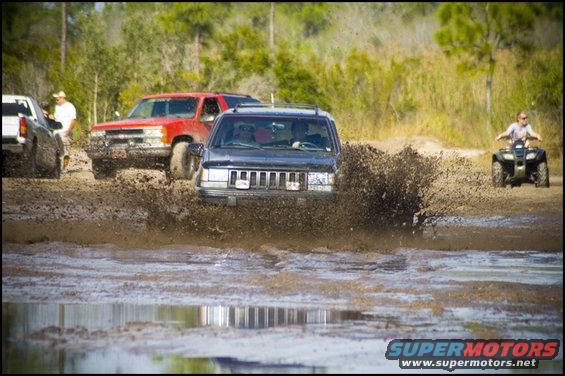 Black Jeep Grand Cherokee driving through mud, front view. Red SUV and white truck behind it