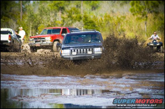 Dark blue SUV splashing through mud, with other vehicles and an ATV in the background