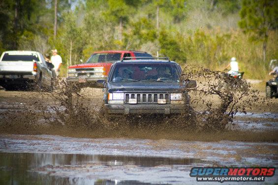 Jeep Cherokee XJ driving through a mud puddle, splashing water. Other trucks and ATVs are visible in the background