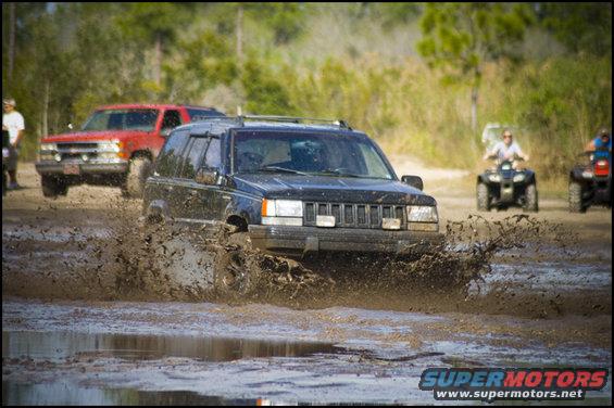 Black Jeep Grand Cherokee splashing through mud. Red truck and ATVs in background