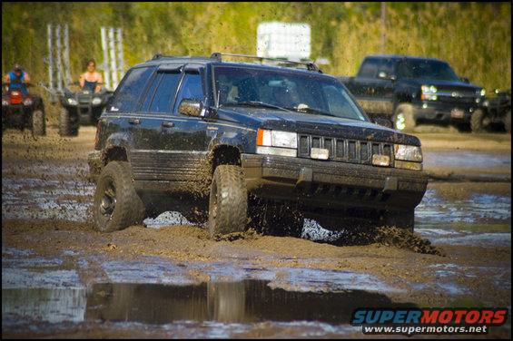 Black Jeep Grand Cherokee driving through mud, with ATVs and a truck in the background