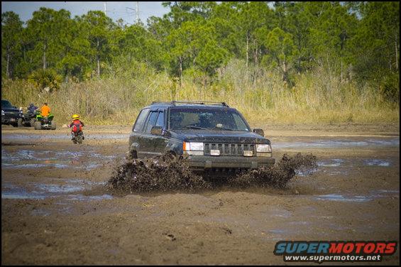 Black Jeep Grand Cherokee splashing through mud, front view. Other vehicles and riders in background