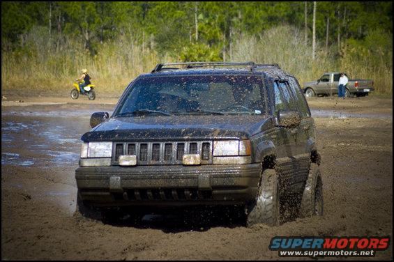 Black Jeep Grand Cherokee stuck in mud, front view. A person on a yellow dirt bike is in the background