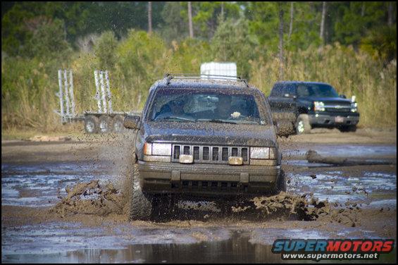 Dark Jeep Grand Cherokee driving through mud, splashing water. Another truck is visible behind it