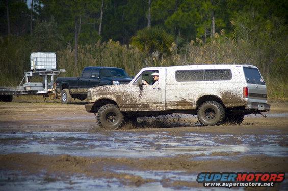 Muddy pickup truck with camper shell driving through mud. Another pickup truck is visible behind it