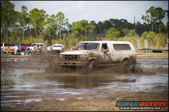 White Ford Bronco splashing through mud pit, other vehicles and spectators in background