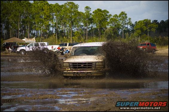 White Ford Econoline van driving through mud, kicking up a lot of spray. Other trucks and a red SUV are visible in the background