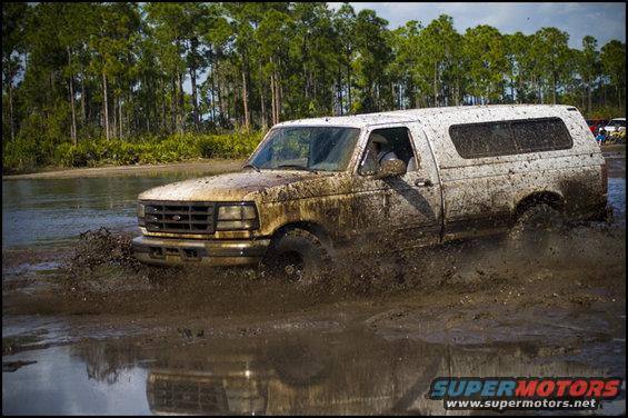 Mud-splattered white Ford F-Series truck with a camper shell driving through a muddy puddle