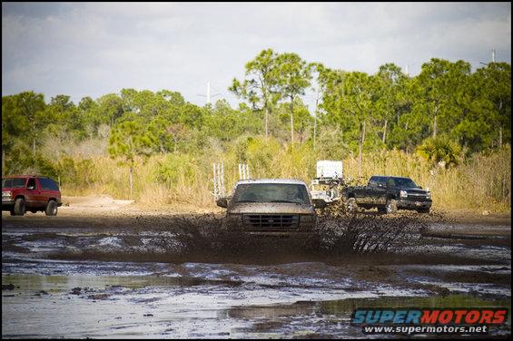 Mud-splattered white Ford E-Series van driving through a mud pit, front view