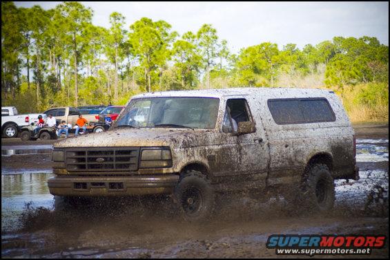 1996 Ford F-150 SUV covered in mud driving through a muddy track with other vehicles in the background