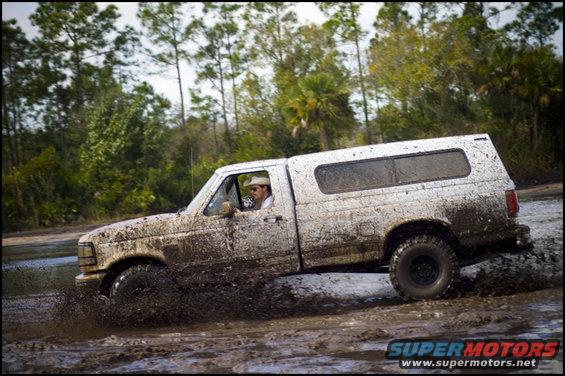 Pickup truck driving through mud, kicking up spray. Driver wears a hat
