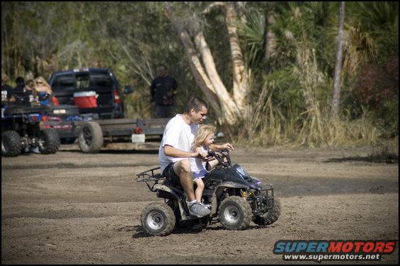 ATV with man and child riding on dirt path, other ATVs and a truck in background