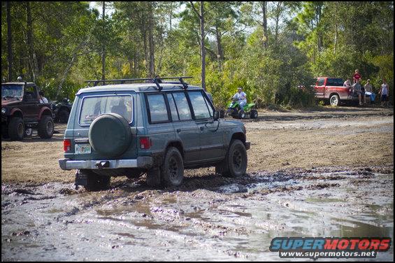 Mitsubishi Pajero driving through mud, red pickup truck and ATV in background