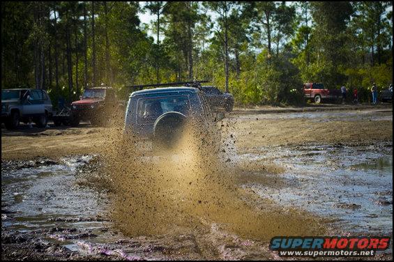 Several off-road vehicles driving through a muddy area, kicking up large amounts of mud and water. Trees and a clear sky are in the background