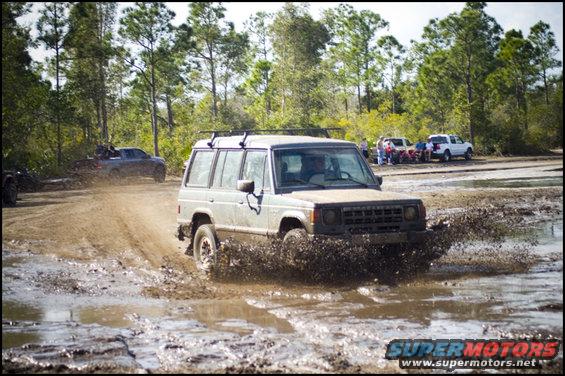 Gray SUV splashing mud on a dirt track, with trees and other vehicles in the background
