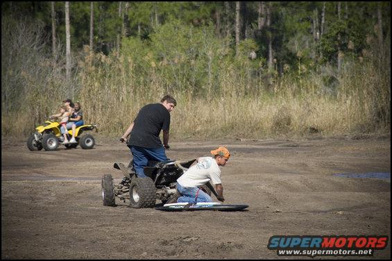 Two ATVs with people riding them in a dirt area with trees and tall grass in the background. One ATV is yellow and has three people on it. The other ATV is black and has two people, one driving and one kneeling