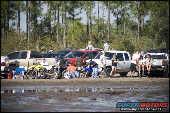People gathered around dirt bikes and pickup trucks in a muddy outdoor area with trees in the background