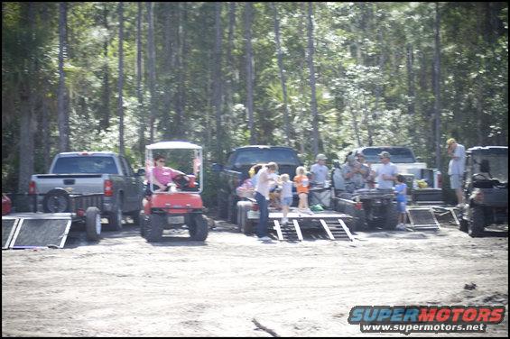 Several trucks, a golf cart, and ATVs are parked in a wooded area with people around