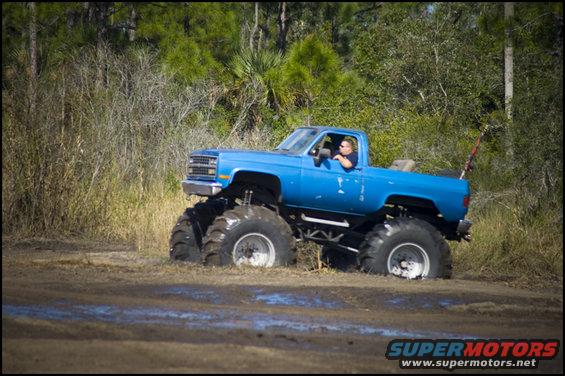 Blue monster truck with large tires stuck in mud, driver visible. Body style suggests 1980s