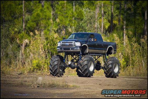 Black monster truck with large tires parked on dirt road, trees in background