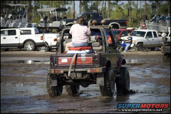 Suzuki Samurai driving through mud with a person in the driver's seat. Other trucks and boats are in the background