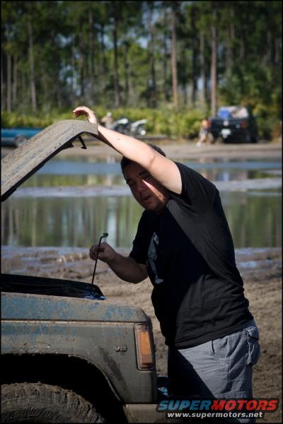 Man checking engine of a muddy blue SUV with V6 badge. Other vehicles and trees in background