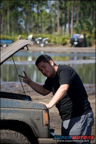 Guy making peace sign by open hood of muddy truck. Other vehicles and trees in background