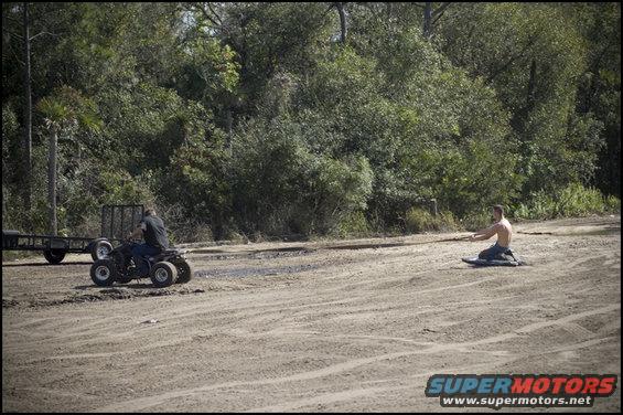 Man on ATV towing a trailer, another man on the ground in a dirt lot