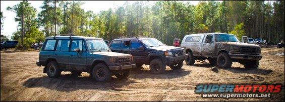 Three SUVs parked in a dirt lot with trees in the background. The SUV on the right is a white pickup truck with a cap