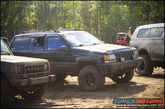 Mud-covered black Jeep Grand Cherokee parked on dirt, front-left view