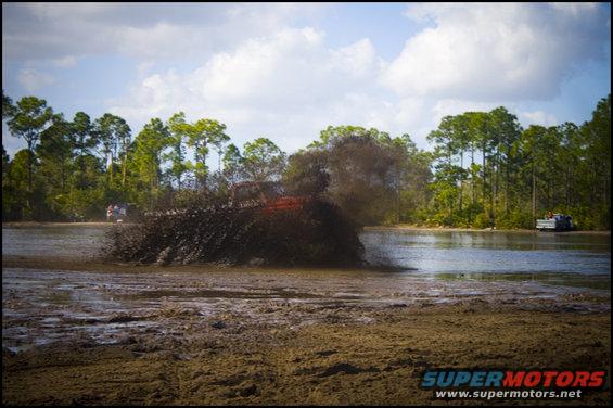 Off-road vehicle driving through mud pit, spraying water and mud. Another vehicle is visible in the background