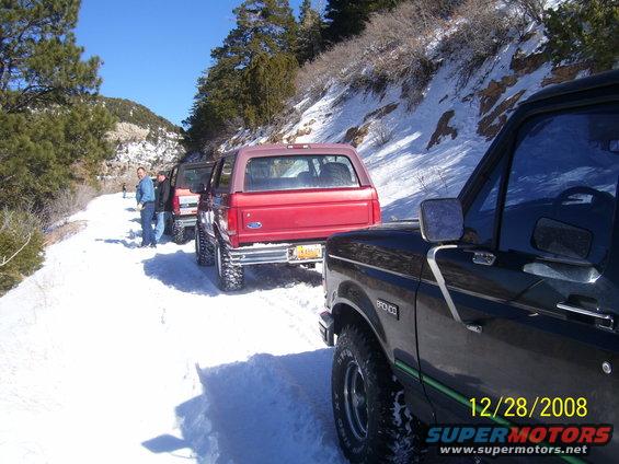 Several Ford Broncos lined up on a snow-covered road with trees and hills in the background. One Bronco is black, another is red
