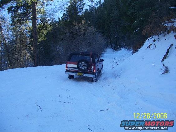 1993 Ford Bronco driving on a snow-covered road in a wooded area. Spare tire mounted on the rear