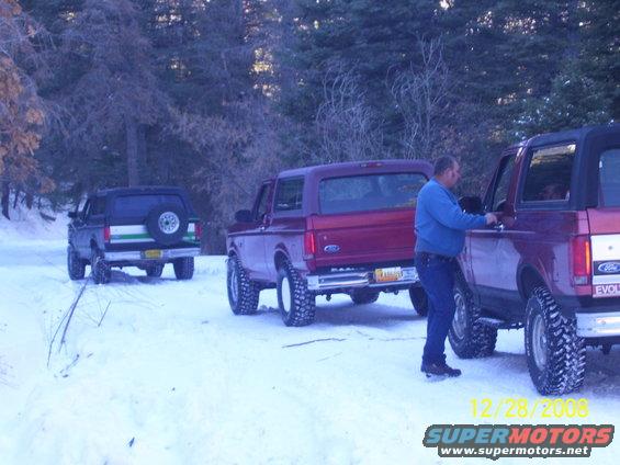 Three Ford Broncos parked on a snowy trail in the woods. One person is standing by the driver's door of the closest Bronco