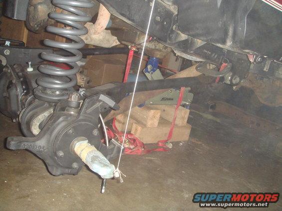 Close-up of a vehicle's front axle assembly with coil spring and brake components. Wood blocks and straps are visible